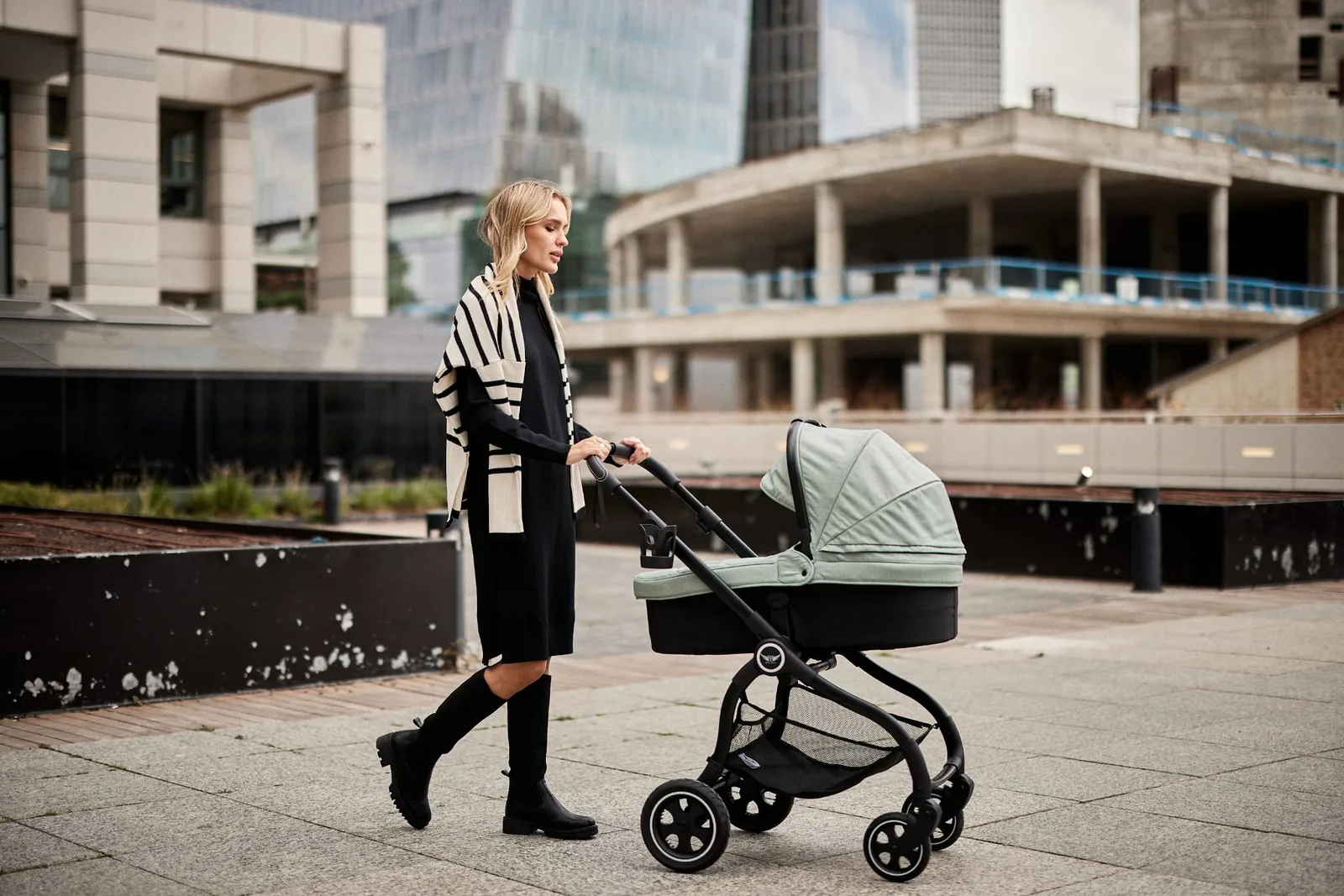 A woman in a black dress pushes a Baby Boss stroller with a mint fabric kit, city towers behind her